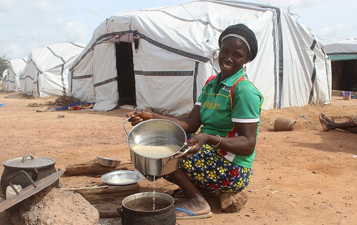 Lady with food in a pot crouching in front of a large while tent with a curved roof known as a Sahelian tent