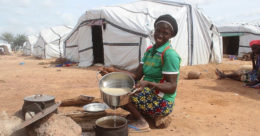 Lady with food in a pot crouching in front of a large while tent with a curved roof known as a Sahelian tent