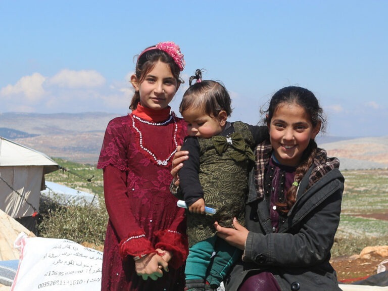Three children in winter clothing standing outside a tent against the landscape in Syria